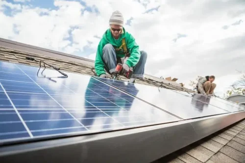 Person in green jacket squatting next to solar panels on a roof