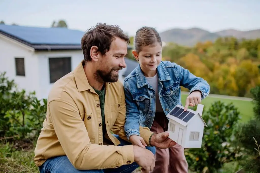 Father and daughter kneeling, holding a mini house with solar panels, pointing at it and smiling.