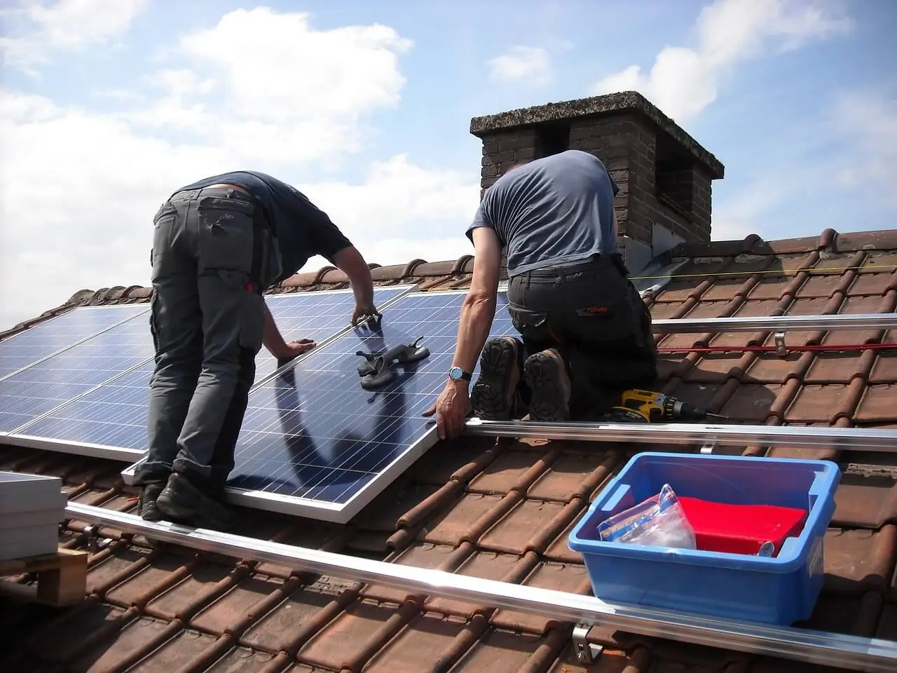 Two people in t-shirts installing solar panels on roof with chimney in background